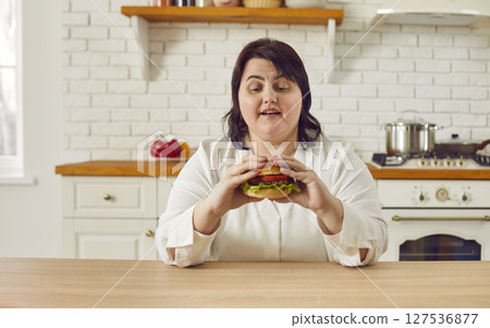 Overweight young woman eating hamburger while sitting at table in kitchen 127536877
