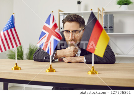 Thoughtful young man sitting at desk in office looking at USA, Great Britain and Germany flags 127536891