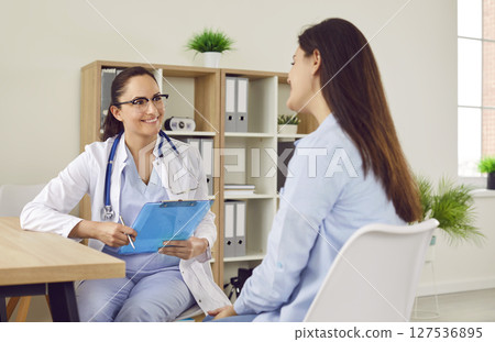 Smiling friendly doctor sitting at the desk examining young pregnant woman in medical clinic. 127536895