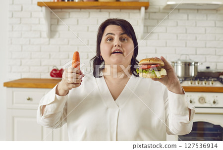 Young overweight plus size fat woman holding a burger in one hand and a carrot in other. Young overweight plus size fat woman holding a burger in one hand and a carrot in other. 127536914