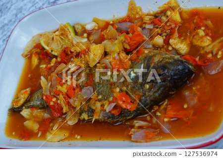 Pecak Ikan Mujair or Fried tilapia doused in a vibrant, spicy, and sour Indonesian chili relish with shallots and lime, served on a white plate. 127536974