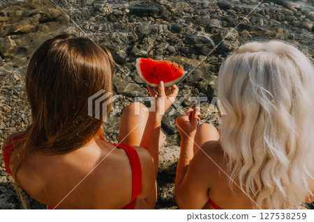 Watermelon Beach Summer: Two women enjoy refreshing watermelon slice on rocky seashore during sunny day. Watermelon Beach Summer: Two women enjoy refreshing watermelon slice on rocky seashore during sunny day. 127538259