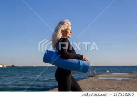 Yoga Mat Woman Beach Exercise - A woman in black athletic wear carries a blue yoga mat on her way to exercise on the beach. Yoga Mat Woman Beach Exercise - A woman in black athletic wear carries a blue yoga mat on her way to exercise on the beach. 127538260