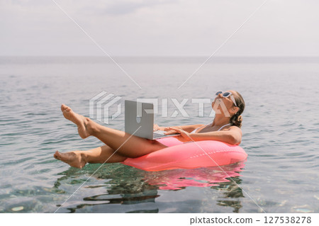 A woman is floating on a pink inflatable raft in the ocean while holding a laptop. Concept of relaxation and leisure, as the woman is enjoying her time in the water while working on her laptop. 127538278