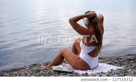 Woman, beach, relaxation: Woman adjusts hair, sitting on beach towel beside lake for relaxation. 127538312