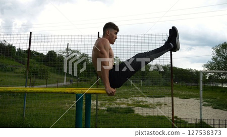 Strong man trains abdominal muscles raising legs on parallel bars at sports ground. Young shirtless sportsman exercising at nature. Sporty athlete working out at nature. Concept of sportive lifestyle 127538373