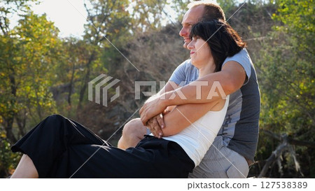 Happy pair hugs during sitting on wooden log on beach. Romantic couple admiring sea landscape at sunny day. Lovers spending time together at resort. Concept of summer vacation or holiday. Slow mo 127538389