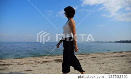 Happy adult woman going on sandy beach near shoreline at sunny day. Beautiful lady walking on seashore with calm waves at background. Concept of summer vacation or holiday. Slow motion Happy adult woman going on sandy beach near shoreline at sunny day. Beautiful lady walking on seashore with calm waves at background. Concept of summer vacation or holiday. Slow motion 127538391