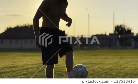 Male feet of professional footballer kicking ball at green field. Legs of young man juggling soccer ball on stadium at sunset. Sportsman practicing tricks outdoor. Freestyle football concept 127538410