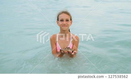 In the calm sea at sunset, a young girl joyfully plays with a starfish as soft waves swirl around 127538562