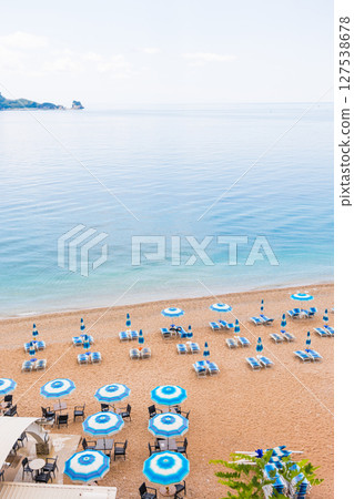 Tranquil morning at a beach with blue umbrellas and loungers. Coastal vacation, summer relaxation and peaceful seaside tourism. Tranquil morning at a beach with blue umbrellas and loungers. Coastal vacation, summer relaxation and peaceful seaside tourism. 127538678