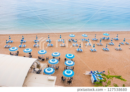 Sunny beach with blue sunbeds and umbrellas near the shore. Summer tourism, seaside relaxation and peaceful coastal vacation. 127538680