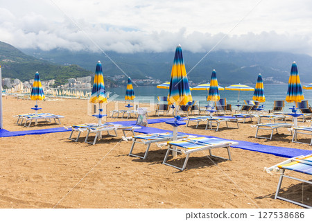 Sunny beach with blue sunbeds and umbrellas near the shore. Summer tourism, seaside relaxation and peaceful coastal vacation. 127538686