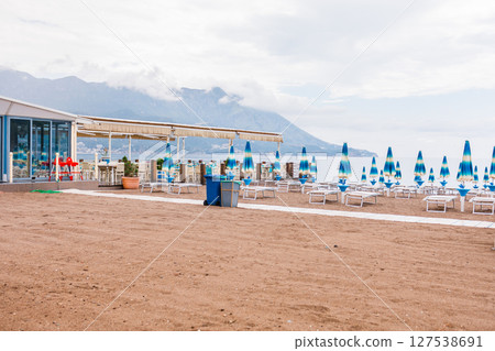 Sunny beach with blue sunbeds and umbrellas near the shore. Summer tourism, seaside relaxation and peaceful coastal vacation. 127538691