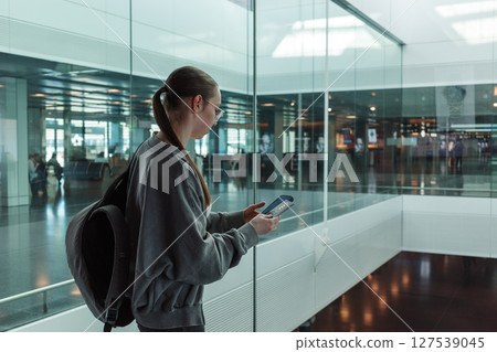 Young woman with a backpack holding a passport and smartphone at a contemporary airport. Concept of international travel, identity verification and global mobility. 127539045