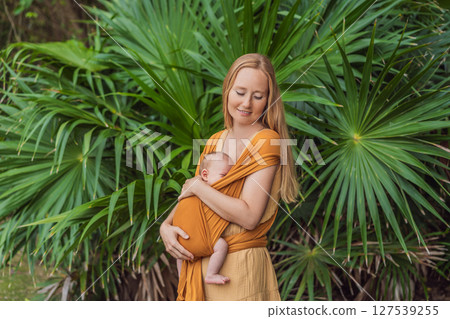 Mother holding her little baby in a yellow sling in the park. Warm and loving family moment. Babywearing, parenting, and mother-child bonding concept Mother holding her little baby in a yellow sling in the park. Warm and loving family moment. Babywearing, parenting, and mother-child bonding concept 127539255