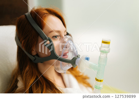 Redhead female patient undergoing respiratory training, comfortably breathing with oxygen mask connected to respiratory device, promoting lung capacity and overall respiratory health, close-up. 127539899