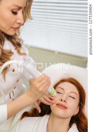 Vertical shot of redhead woman receiving microneedling rejuvenation treatment in clinic of aesthetic medicine lying with closed eyes. Cosmetologist performing needle mesotherapy on female client face. Vertical shot of redhead woman receiving microneedling rejuvenation treatment in clinic of aesthetic medicine lying with closed eyes. Cosmetologist performing needle mesotherapy on female client face. 127539904