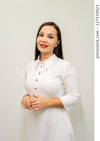 Friendly female doctor wearing white uniform and red lipstick, standing confidently on white isolated background, smiling looking at camera, radiating medical expertise and skincare knowledge. 127539923