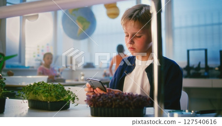 Elementary School Boy Sitting at the Table and Using Smartphone, Growing Experimental Microgreens Elementary School Boy Sitting at the Table and Using Smartphone, Growing Experimental Microgreens 127540616
