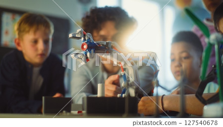 Team of Talented Kids Sitting at Table, Studying Robotic Arm Model with Teacher in Primary School 127540678