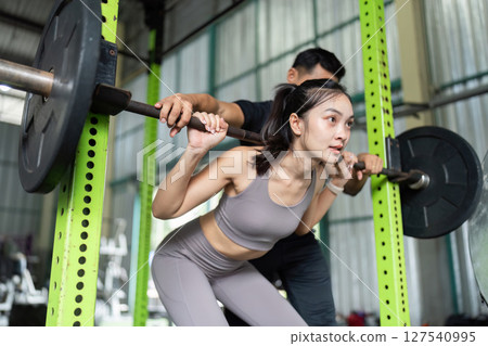 Partner Workout. Woman squatting with support from trainer in gym. Partner Workout. Woman squatting with support from trainer in gym. 127540995