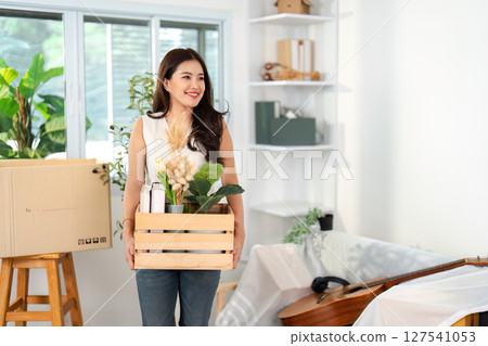 Joyful Moving Day. A woman smiling while holding a crate of plants in her new home. 127541053
