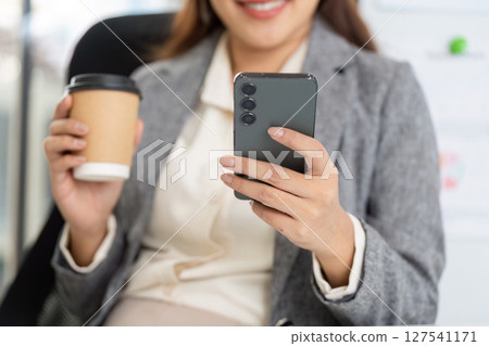 Professional woman checking smartphone while enjoying coffee in office 127541171