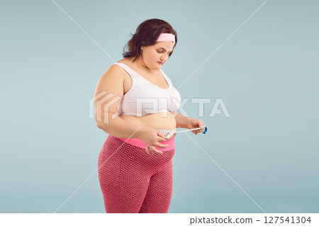 Fat woman wearing sportswear measure waist with measuring tape isolated on blue background. 127541304