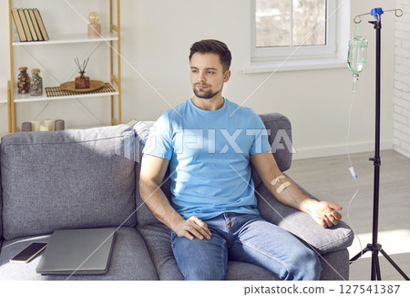Young man receiving IV drip infusion and vitamin therapy sitting on the couch at home. Young man receiving IV drip infusion and vitamin therapy sitting on the couch at home. 127541387