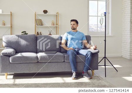 Young man receiving IV drip infusion and vitamin therapy sitting on the couch at home. 127541414