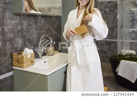 Portrait of young cheerful woman brushing hair with hairbrush standing in modern bathroom interior 127541424