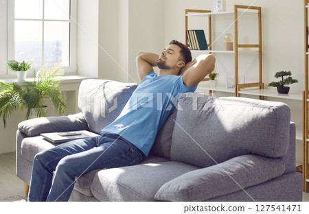 Man relaxing on a comfortable couch, with his hands behind his head and a laptop beside Man relaxing on a comfortable couch, with his hands behind his head and a laptop beside 127541471