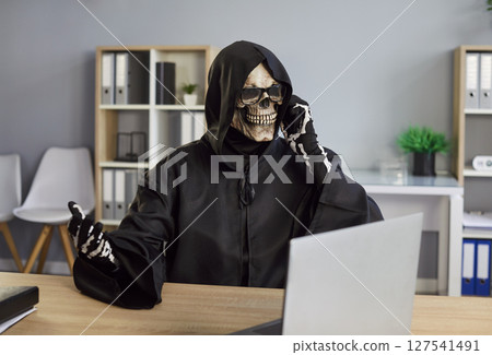 Man in death costume with black hood and skeleton mask sitting in office talking by mobile phone Man in death costume with black hood and skeleton mask sitting in office talking by mobile phone 127541491
