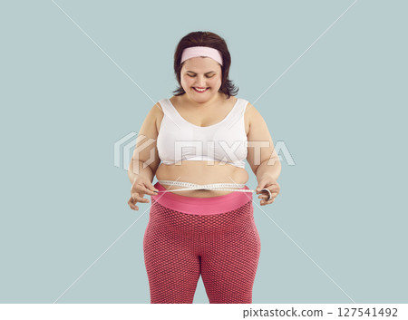 Fat woman wearing sportswear measure waist with measuring tape isolated on blue background. 127541492