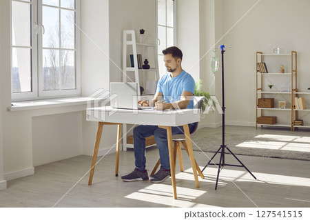 Young man receiving intravenous vitamin therapy while working with laptop at his workplace Young man receiving intravenous vitamin therapy while working with laptop at his workplace 127541515