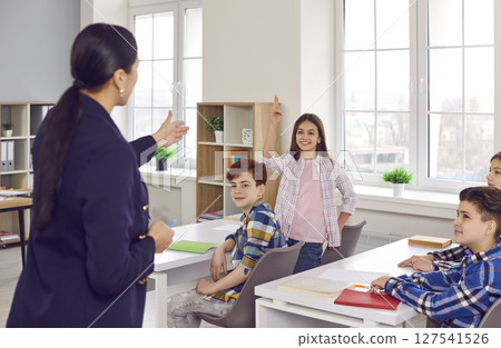 Schoolgirl answering on a lesson standing at the desk to a female teacher in classroom. 127541526