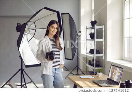 Portrait of happy young woman photographer in studio with laptop and lighting equipment 127541612