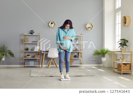 Portrait of a young pregnant woman measuring her weight standing on a scales at home. Portrait of a young pregnant woman measuring her weight standing on a scales at home. 127541643