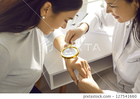 Female doctor examining hand skin of a young woman patient using magnifying glass. Female doctor examining hand skin of a young woman patient using magnifying glass. 127541660