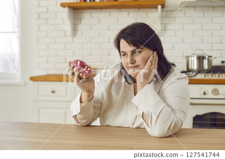 Pensive overweight young woman hesitating to eat donut 127541744