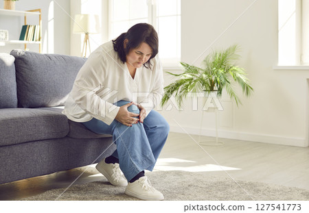 Young overweight woman holding a knee suffering from osteoarthritis sitting on sofa at home. Young overweight woman holding a knee suffering from osteoarthritis sitting on sofa at home. 127541773