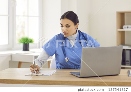 Young woman doctor sitting at desk, working on laptop computer, and writing notes Young woman doctor sitting at desk, working on laptop computer, and writing notes 127541817