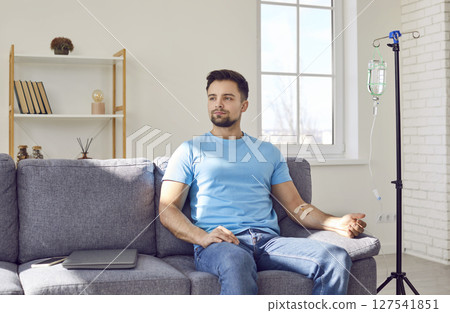 Young man sitting on the sofa at home and getting an intravenous IV line vitamin infusion Young man sitting on the sofa at home and getting an intravenous IV line vitamin infusion 127541851
