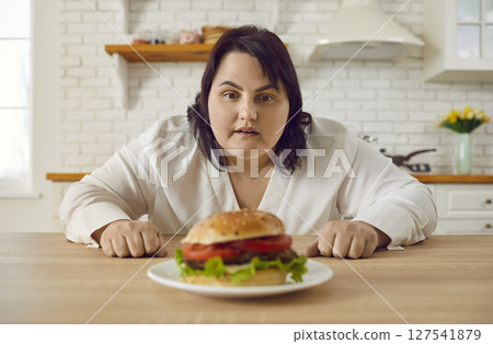 Young fat excited woman sitting at the table in kitchen looking at the plate with big burger. 127541879