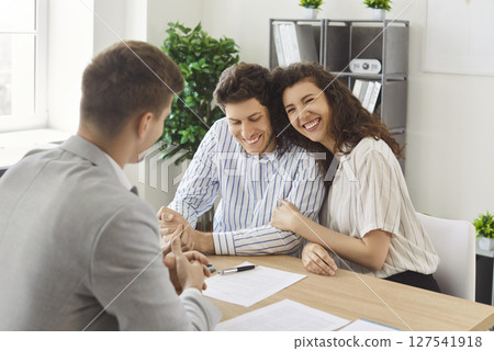 Happy young couple hugging and smiling during meeting with realtor, celebrating property purchase. 127541918
