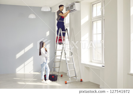 Young woman homeowner watching the electrician man maintaining modern air conditioner indoors. 127541972