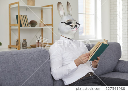 Young man in funny rabbit mask and eyeglasses sitting on couch and reading novel Young man in funny rabbit mask and eyeglasses sitting on couch and reading novel 127541988