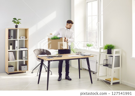 Fired male office worker standing by his desk and packing his stuff in cardboard box 127541991