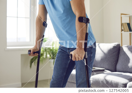 Injured or disabled young male patient walking with crutches in living room at home 127542005